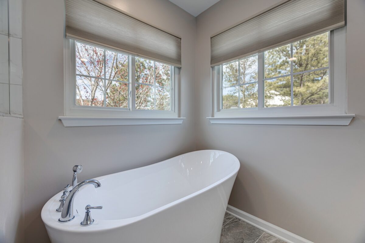 Freestanding soaking tub beneath two windows with gray tile floor